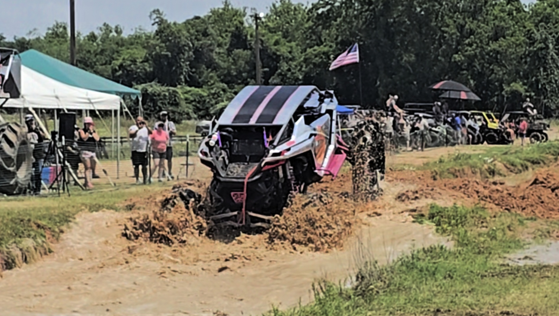 Pink and black side-by-side splashing through a muddy race course beside spectators